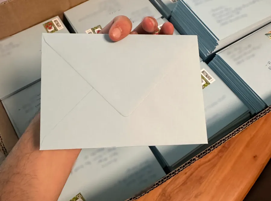 Eine Hand hält einen zugeklebten blauen Umschlag über Kisten voller versandfertiger Briefe